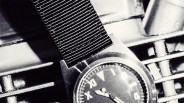 Black and white close-up of a wristwatch with a fabric strap resting on a metal grate.