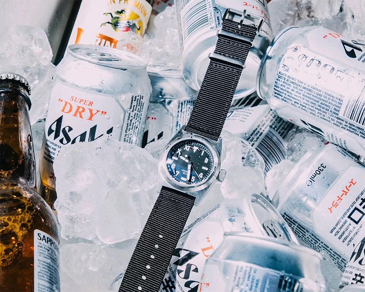 Black strap wristwatch with a black dial placed on ice among white Asahi beer cans and a brown bottle.