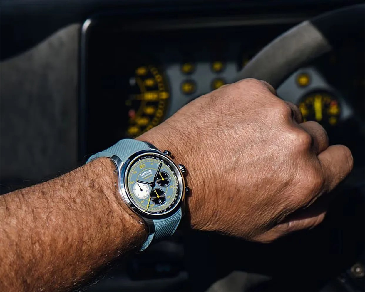 Wristwatch with a blue textured strap and a multi-colored chronograph dial worn on a hand gripping a steering wheel.