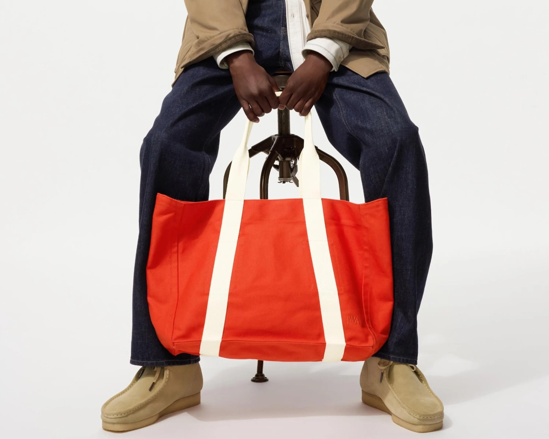 Bright orange canvas tote bag with long white handles held by a person sitting on a stool.