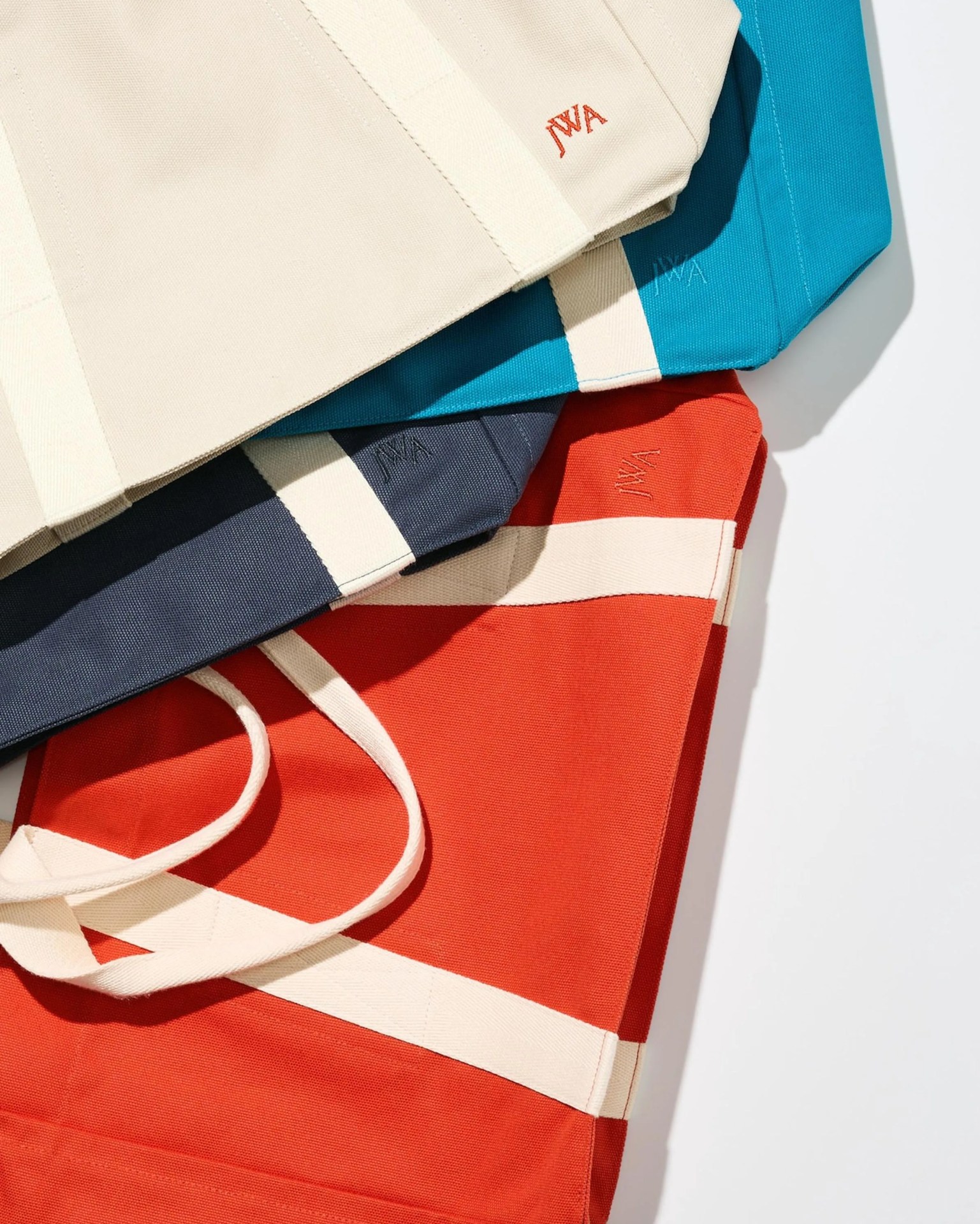 Stack of four canvas tote bags in cream, teal, navy, and red, each with contrasting white straps and a small embroidered logo.