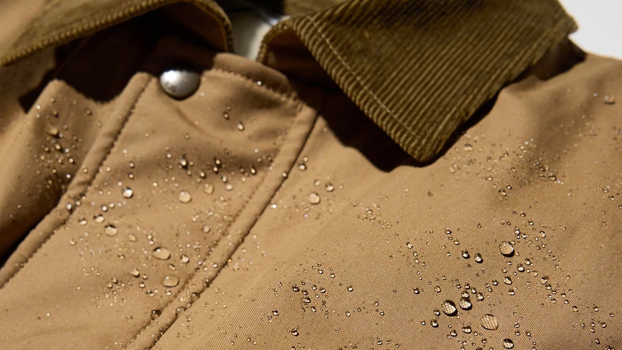 Close-up of a tan water-resistant jacket with water droplets on the fabric and a brown corduroy collar.