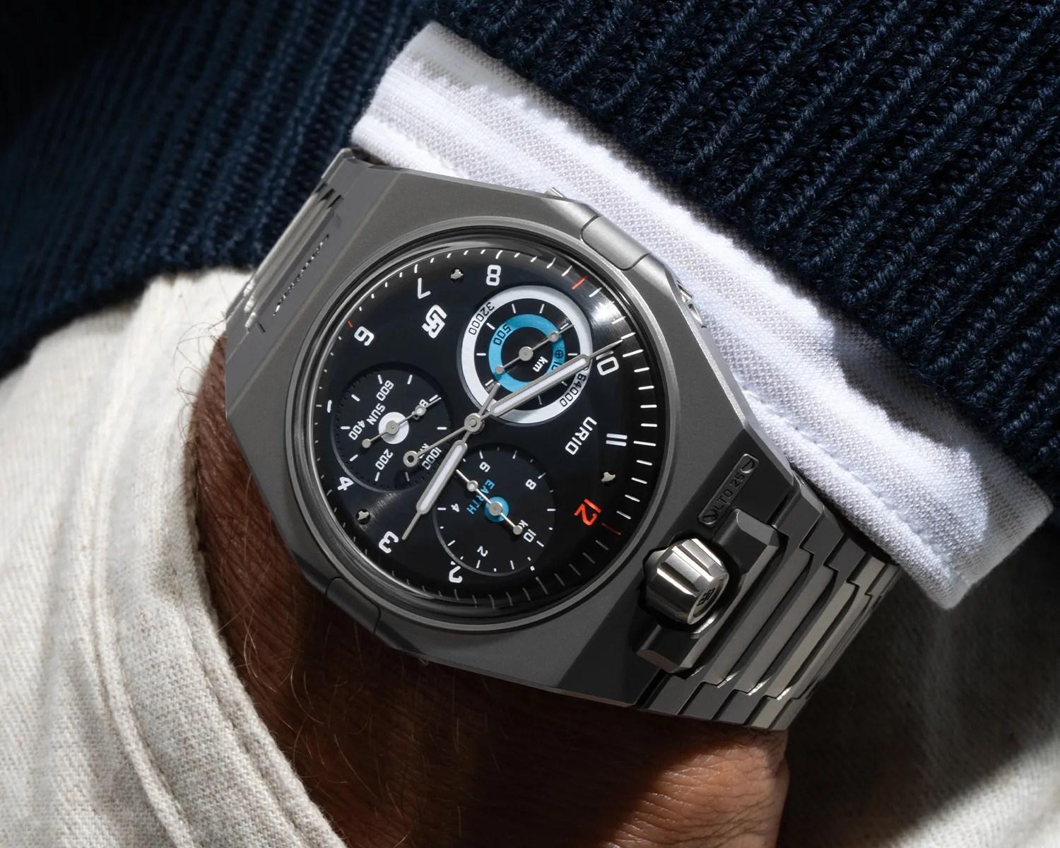 Matte gray metal wristwatch with black dial, blue and white subdials, and integrated metal bracelet on a wrist.