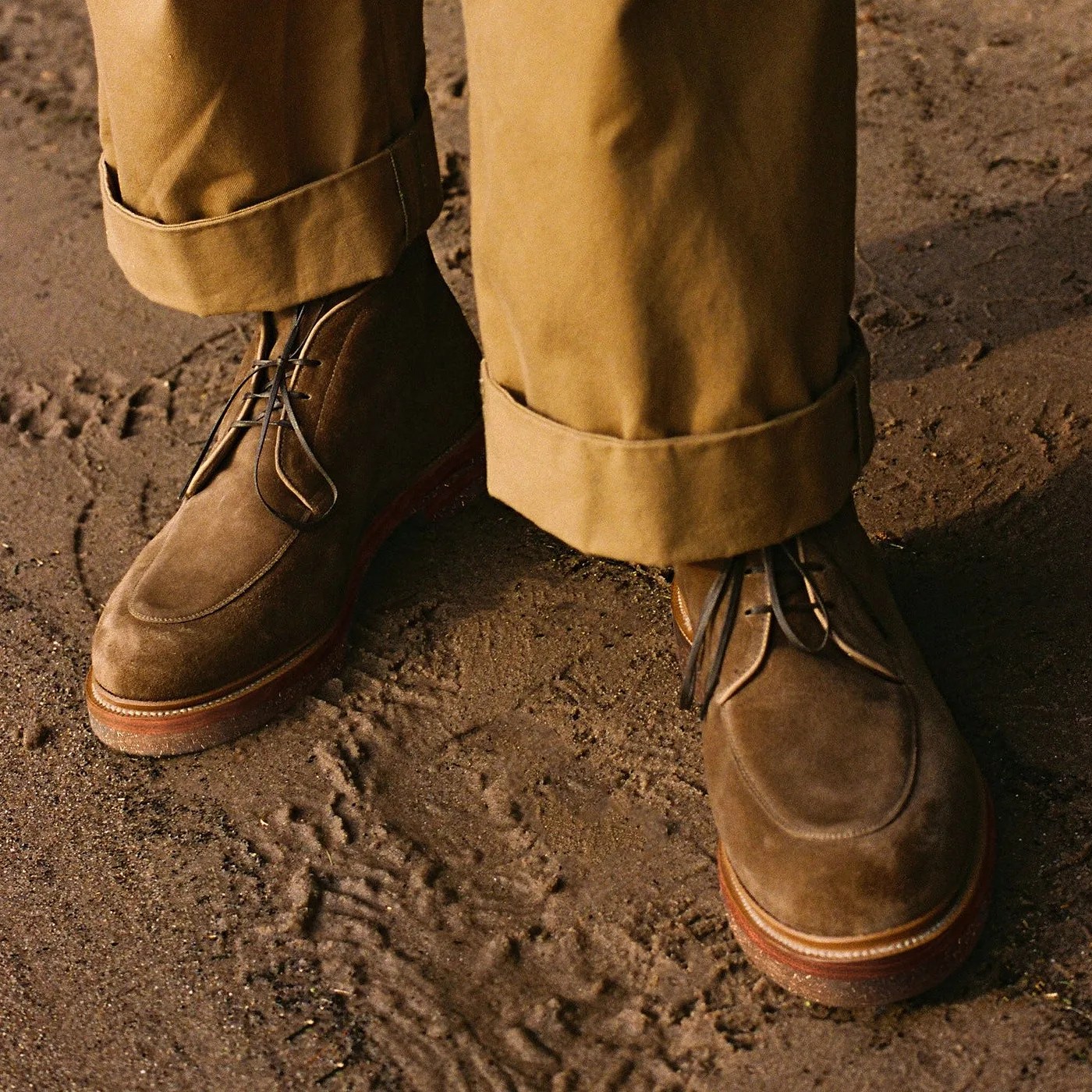Brown suede lace-up boots worn with rolled-up tan pants on dirt ground.
