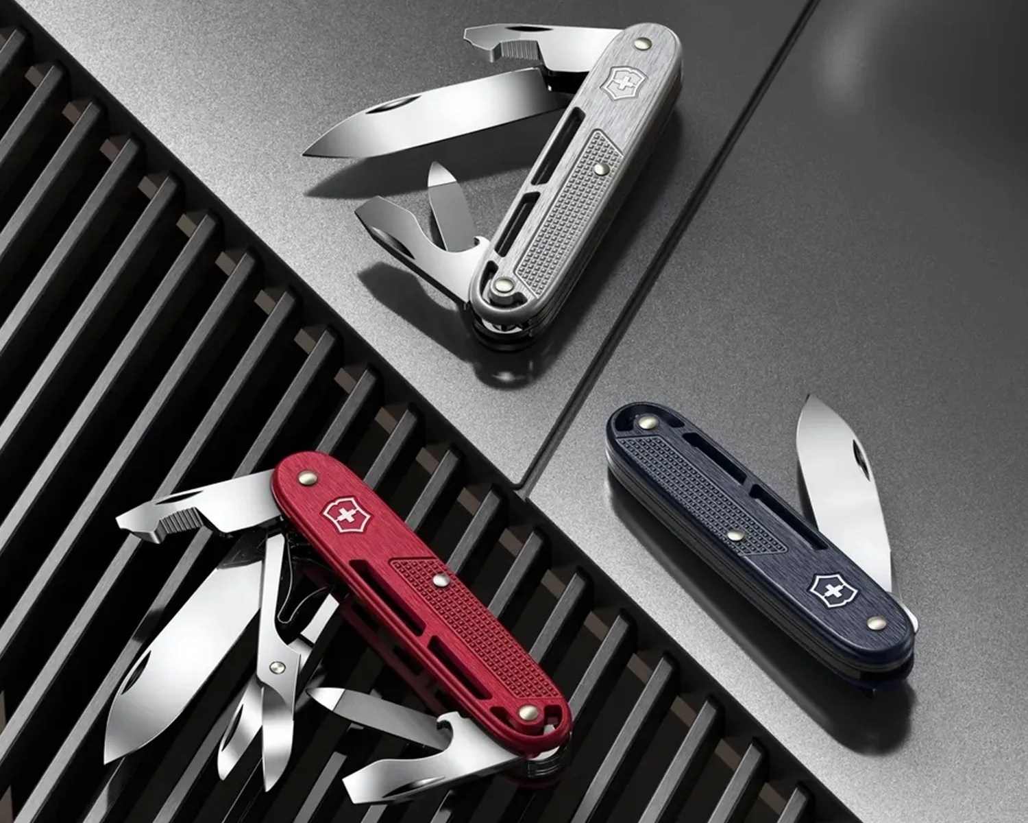 Three Victorinox Swiss Army knives in silver, red, and navy blue with various tools extended on a dark textured surface.