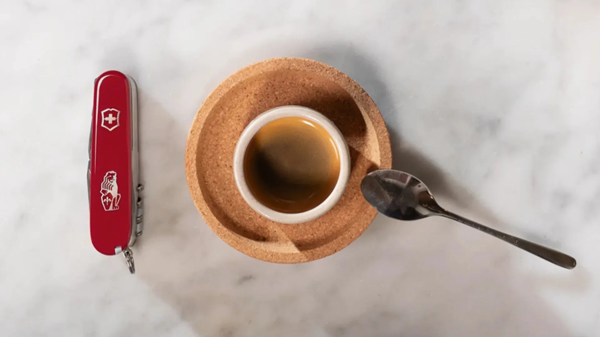 Espresso cup on a cork saucer with a spoon to the right and a red Swiss Army knife to the left on a marble surface.