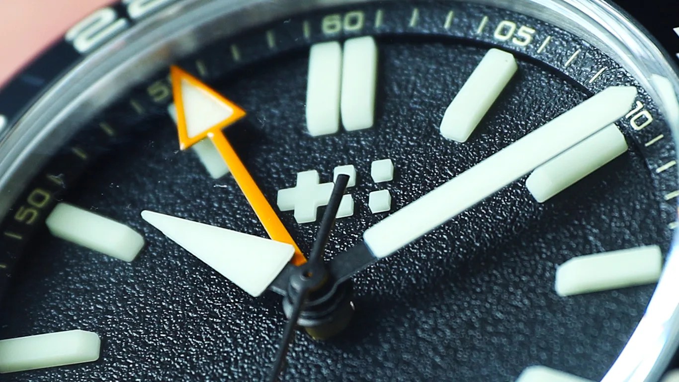 Close-up of a black textured watch face with large white hour markers and white and orange hands.