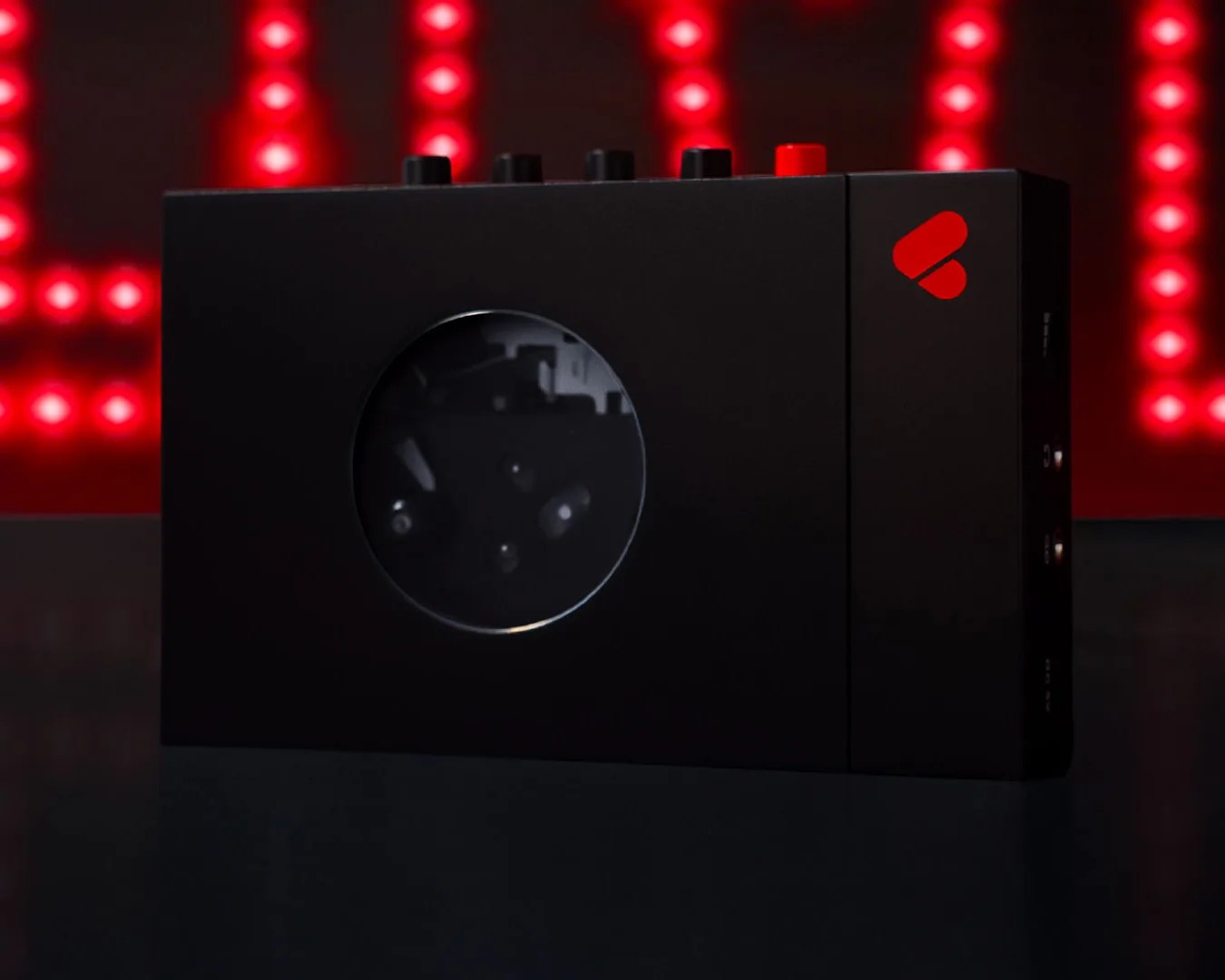 Black rectangular electronic device with a circular window and red logo on the right side, set against a background of red lights.