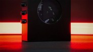 Black rectangular device with three control knobs on the left side and a circular window on the front, illuminated by red light.