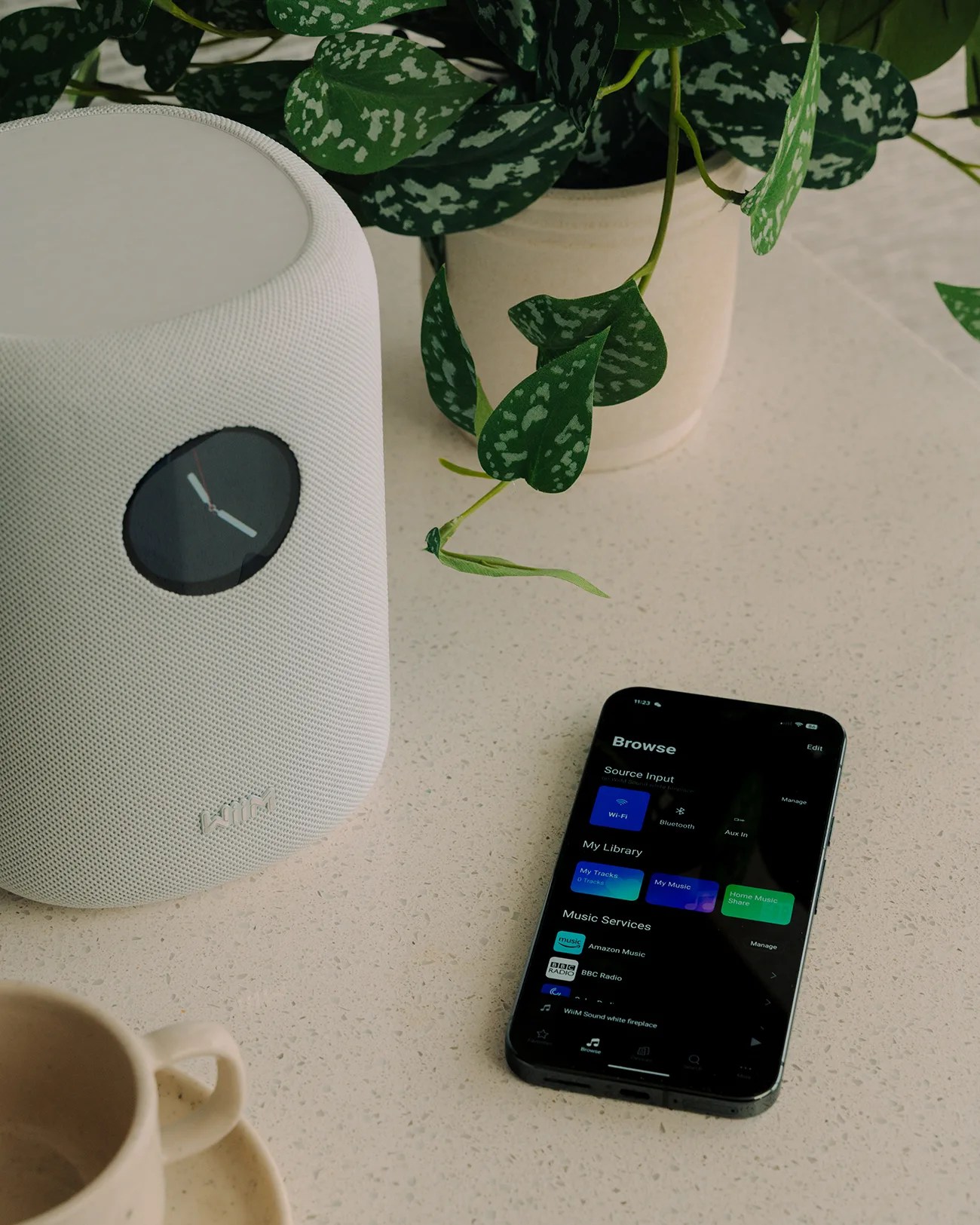 White cylindrical smart speaker with a round clock display next to a smartphone showing a music app on a beige countertop.