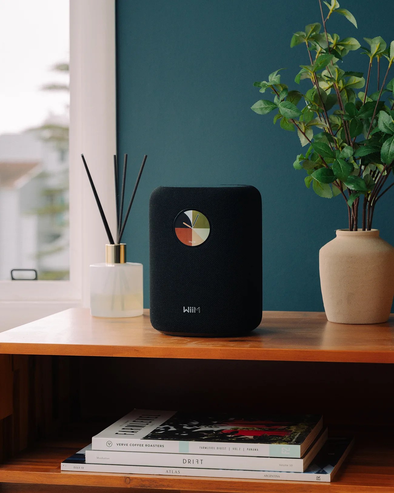 Black rectangular smart speaker with a round multicolor clock display on a wooden table next to a reed diffuser and a beige vase with green leaves.