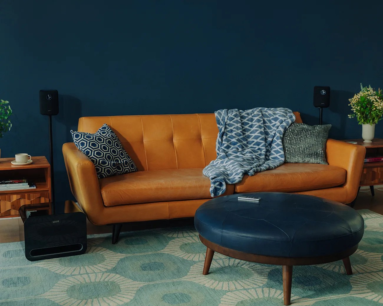 Tan leather sofa with patterned blue and gray pillows and a blue patterned throw, next to a round navy ottoman on a patterned rug.