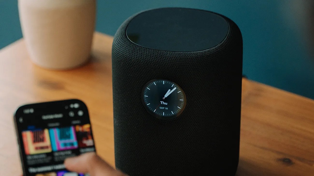 Black cylindrical smart speaker with a round digital clock display showing "Thu SEP 18" on a wooden surface.