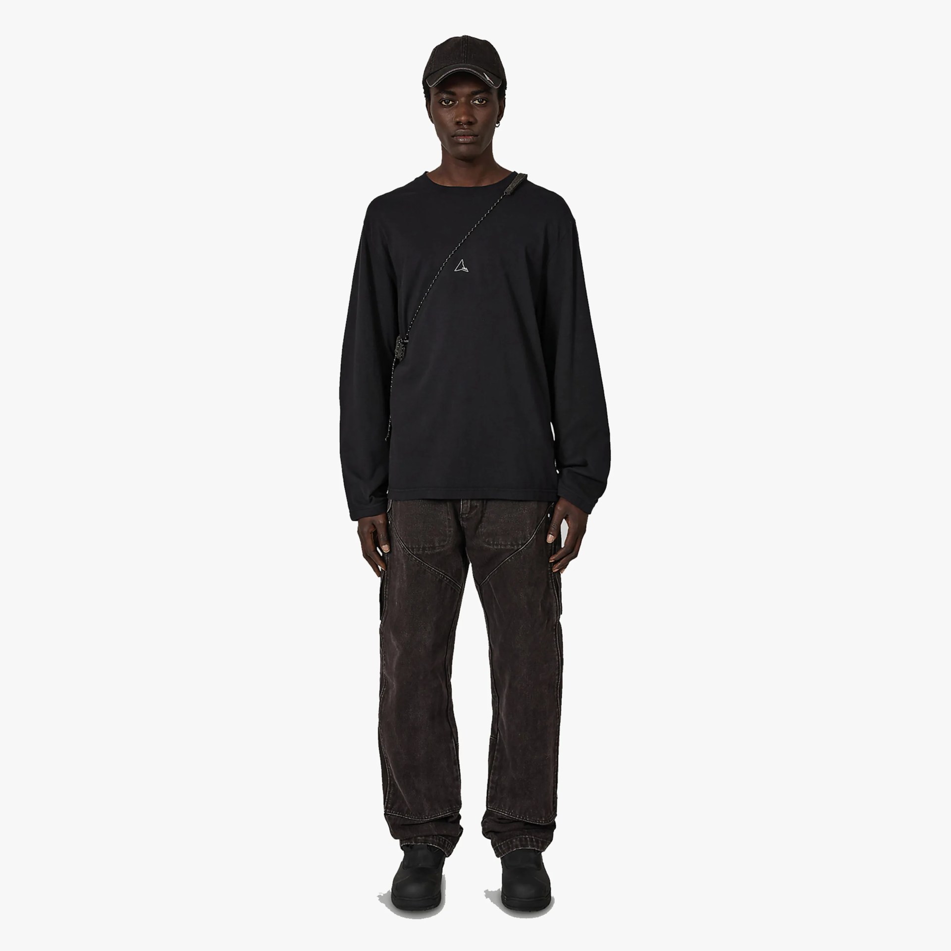 Person wearing a black long-sleeve shirt with a small white logo, dark brown pants, black shoes, and a black cap.