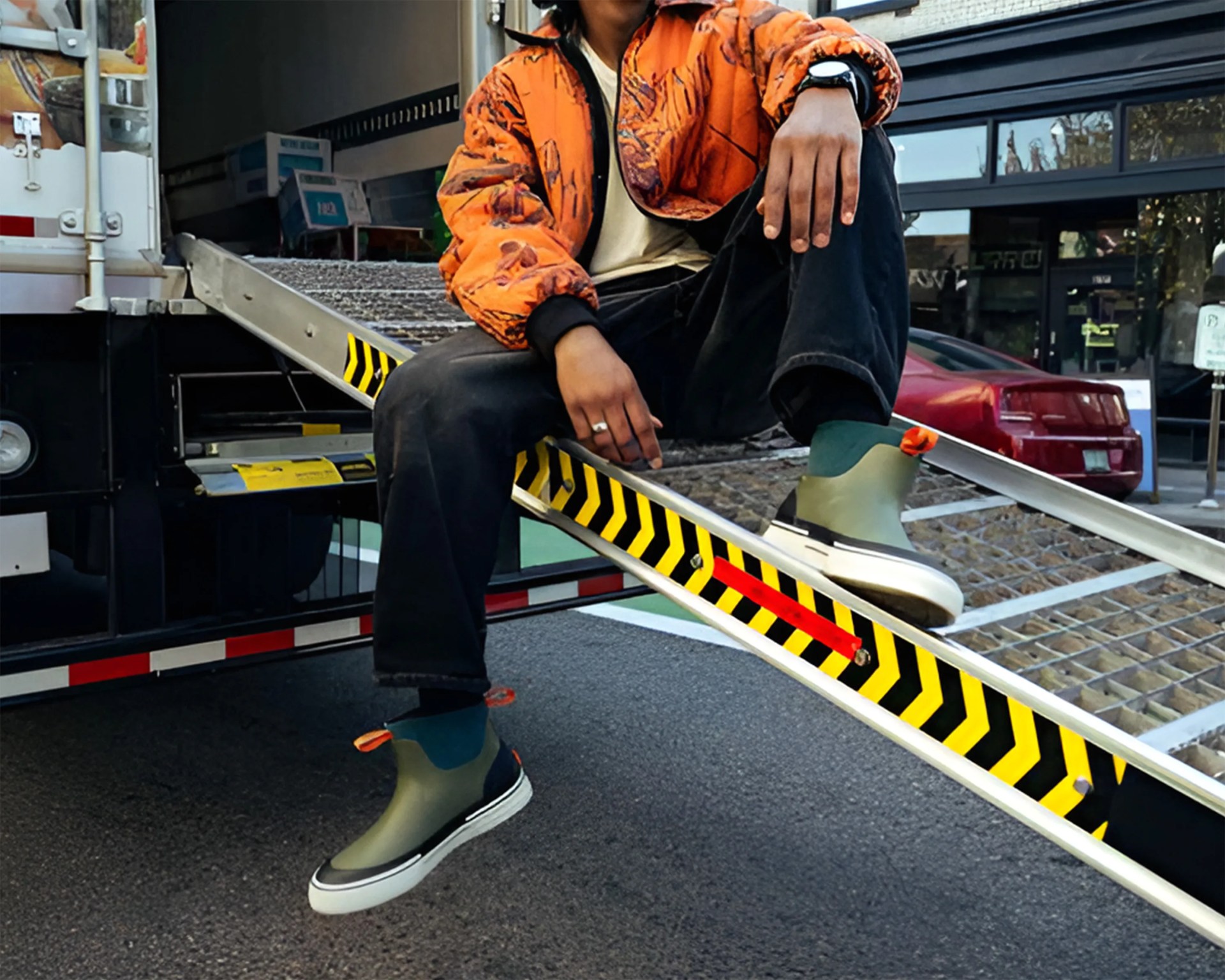 Person wearing green and black slip-on boots with orange pull tabs, black pants, and an orange patterned jacket sitting on a metal ramp.