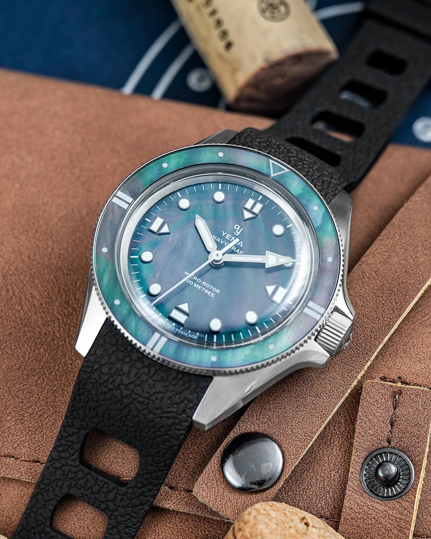 Yema Navyraf wristwatch with blue-green iridescent dial and bezel, black textured strap, on brown fabric.