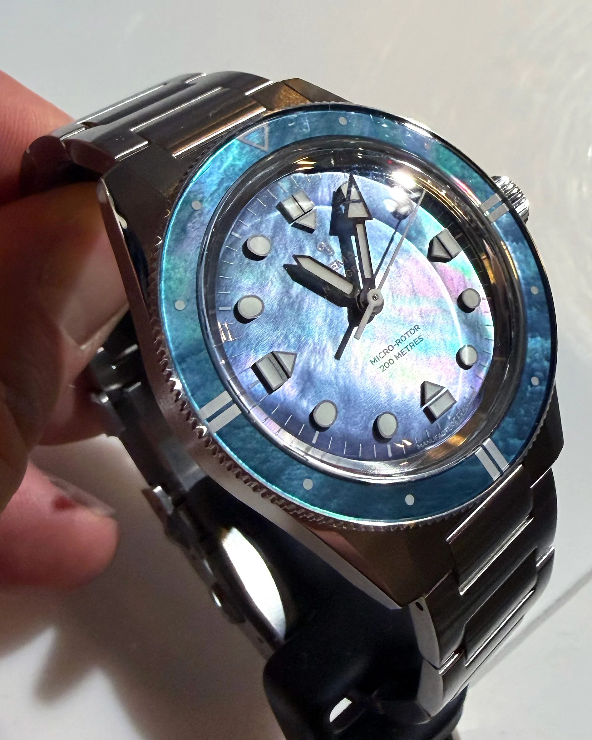 Silver metal wristwatch with a blue iridescent dial and bezel, held in a hand.