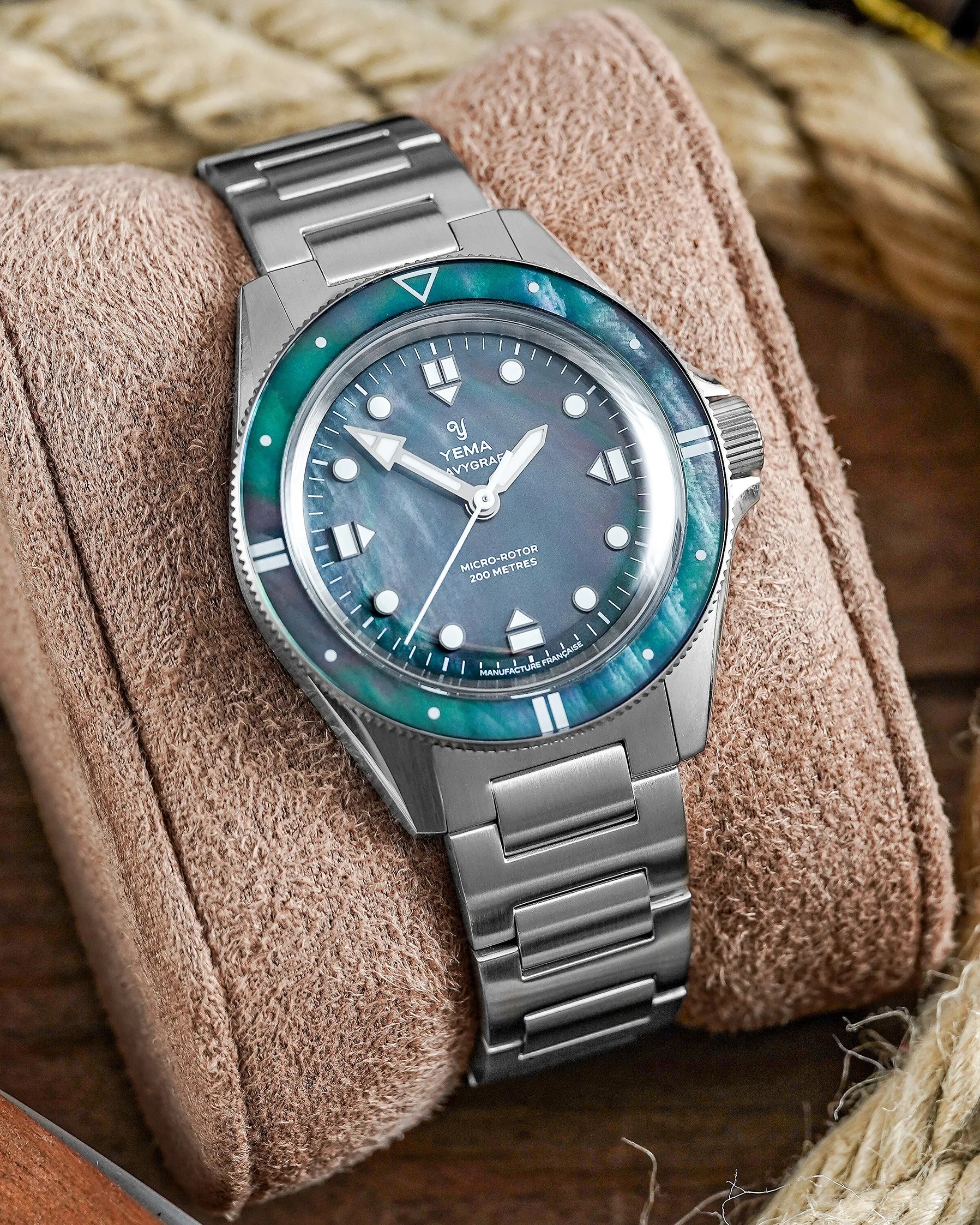 Stainless steel Yema Navygraf wristwatch with blue-green mother-of-pearl bezel and dial on a beige cushion.