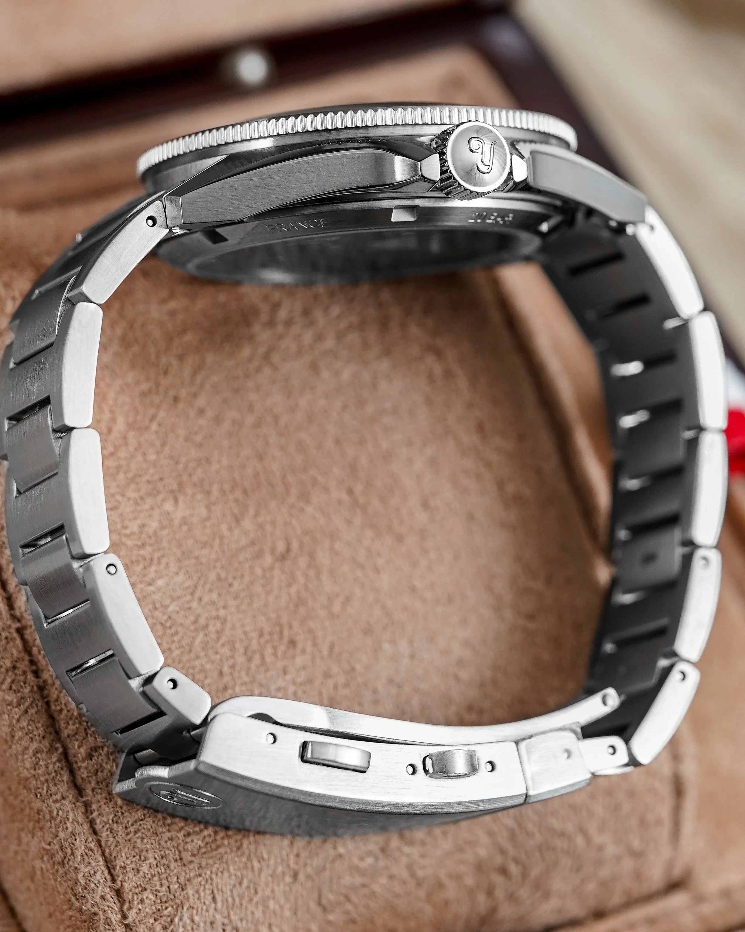 Brushed stainless steel wristwatch with linked bracelet and a crown engraved with a stylized "Y" on a tan suede surface.