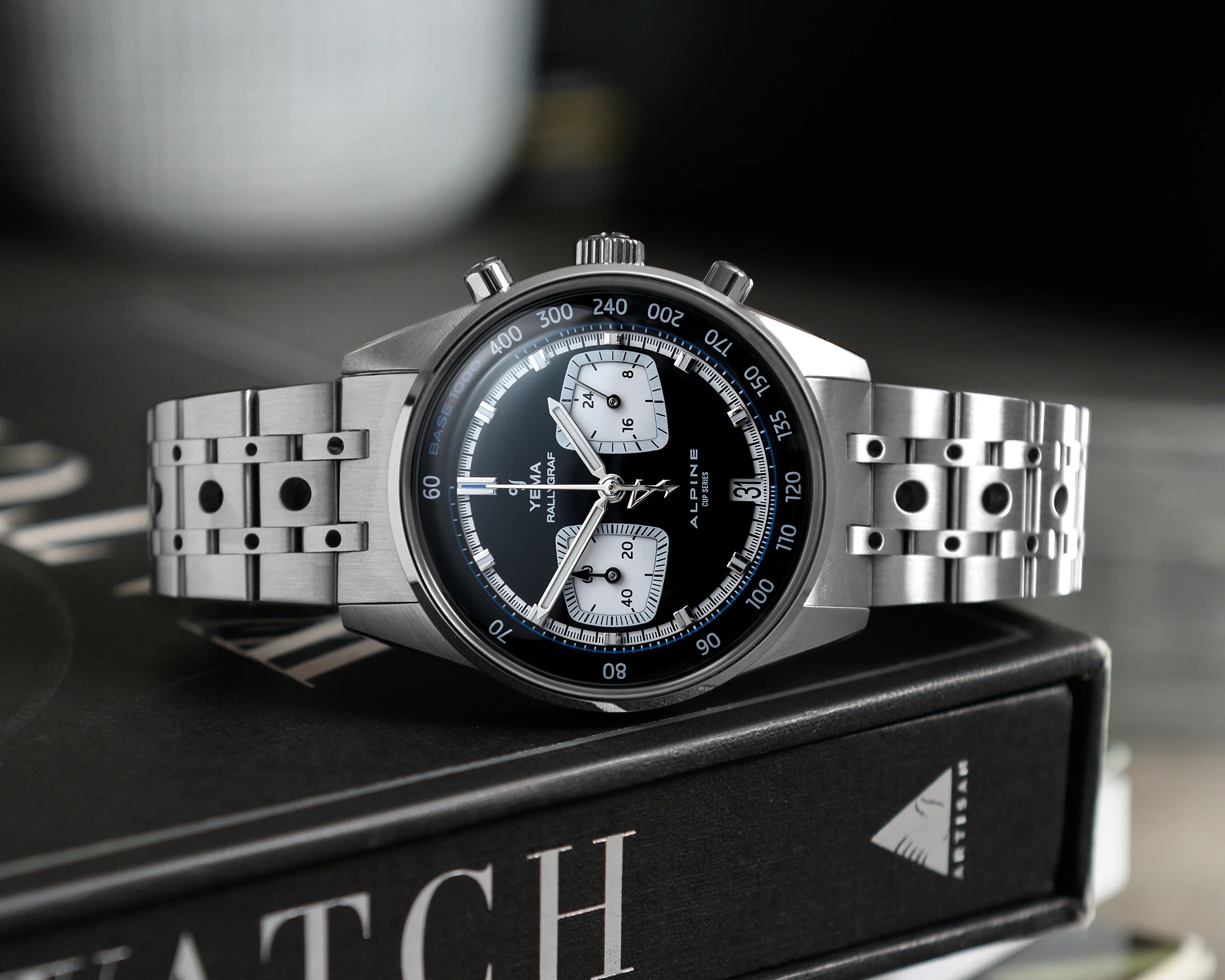 Stainless steel Yema Rallygraf Alpine chronograph watch with black dial and perforated metal bracelet on a book.