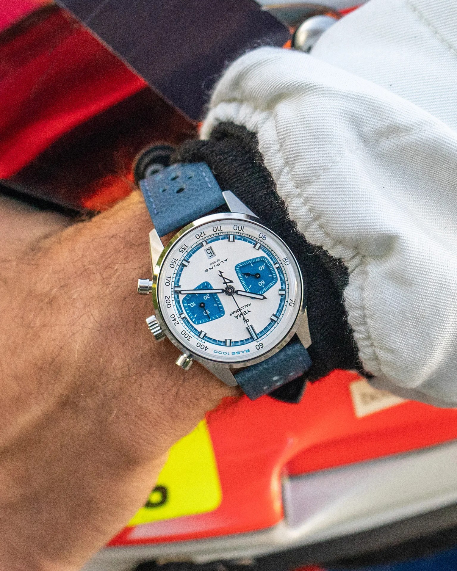 Silver chronograph wristwatch with white dial and blue subdials on a blue perforated strap worn on a wrist.