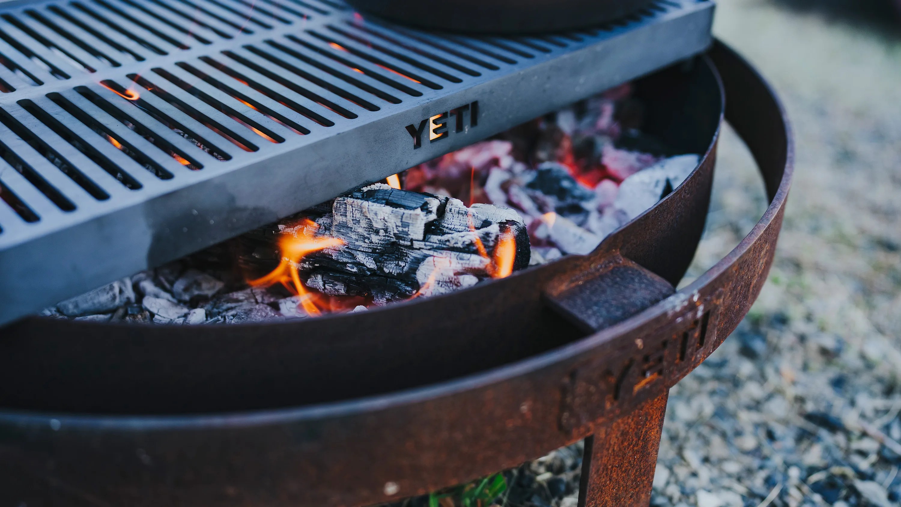 Rusty round metal fire pit with burning charcoal and a black YETI grill grate on top.