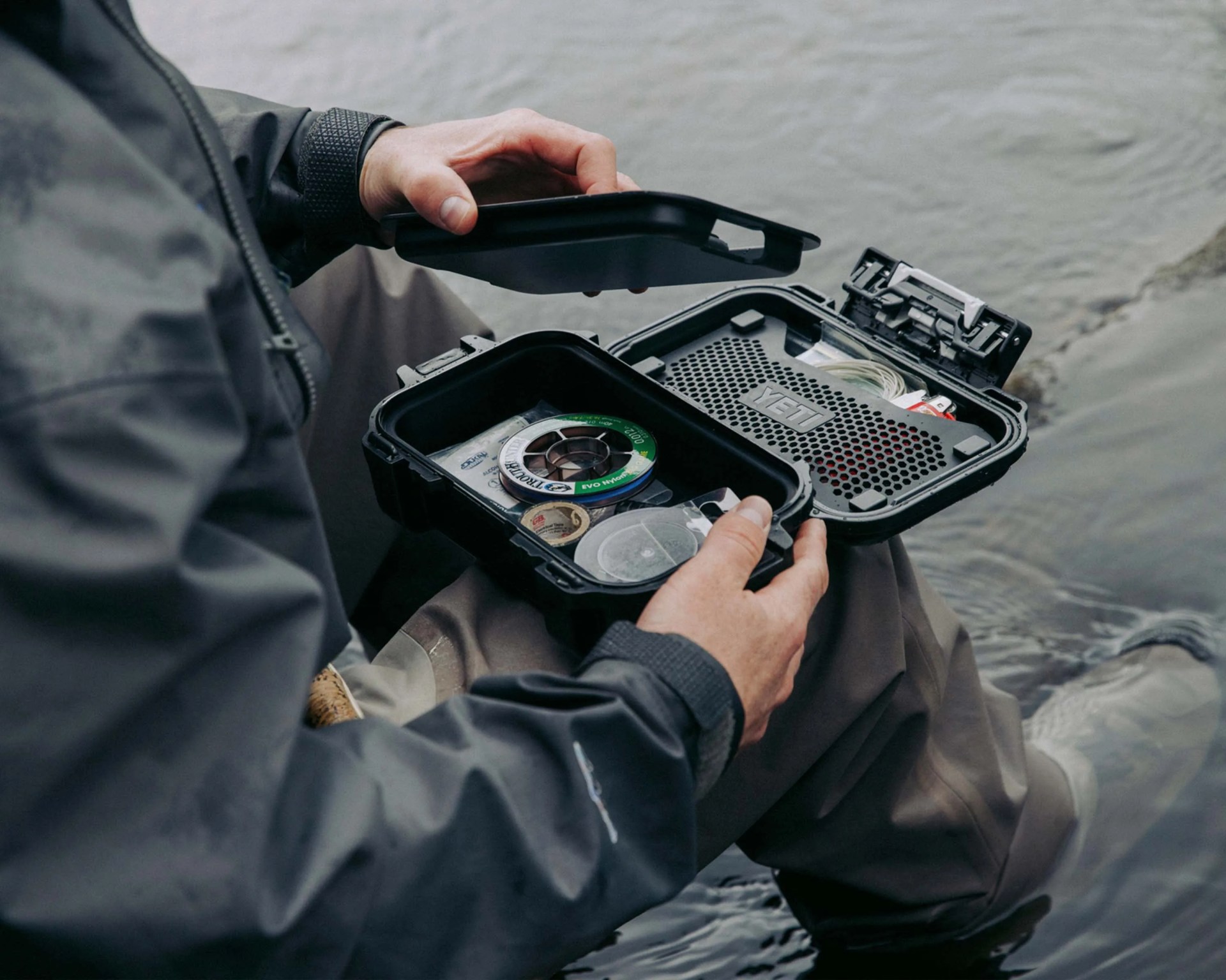 Person holding open a black YETI waterproof case with fishing line and accessories inside near water.