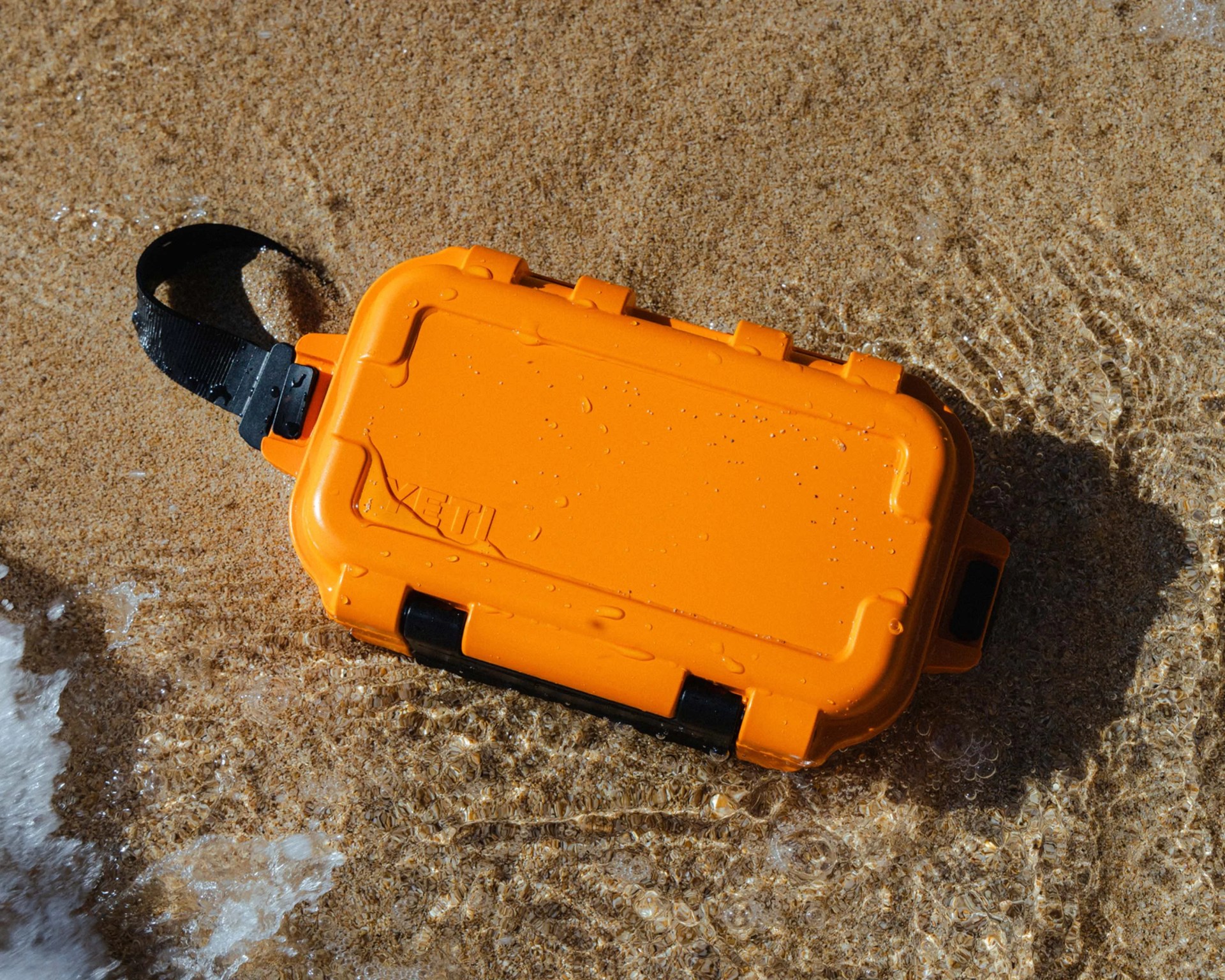 Orange YETI waterproof hard case with black strap resting on wet sandy beach.