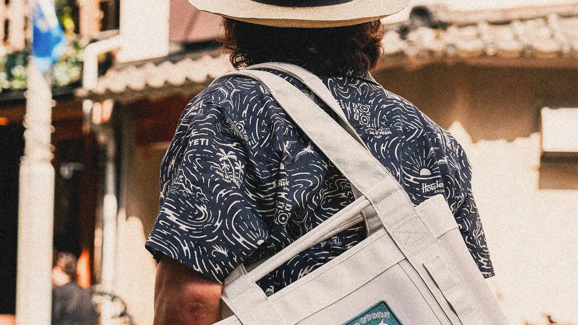 Person wearing a navy patterned shirt with "YETI" and "Howler Bros" logos, carrying a beige canvas bag with a green patch.