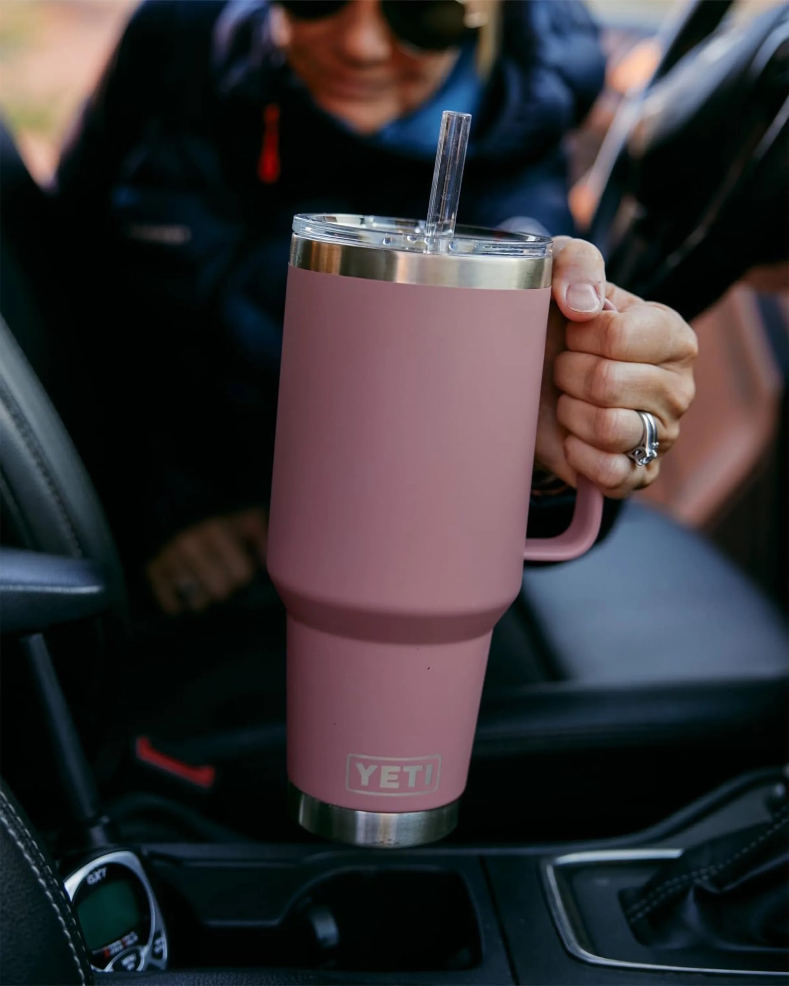 Pink YETI tumbler with clear lid and straw held inside a car.