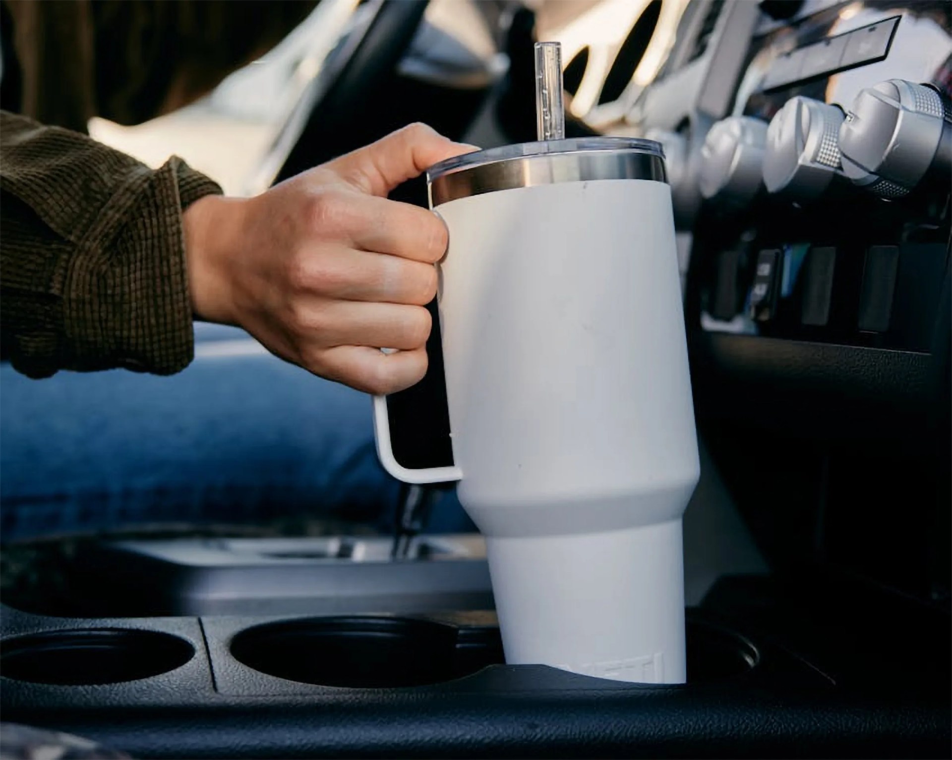 Hand holding a white insulated travel mug with a handle and straw inside a car cup holder.