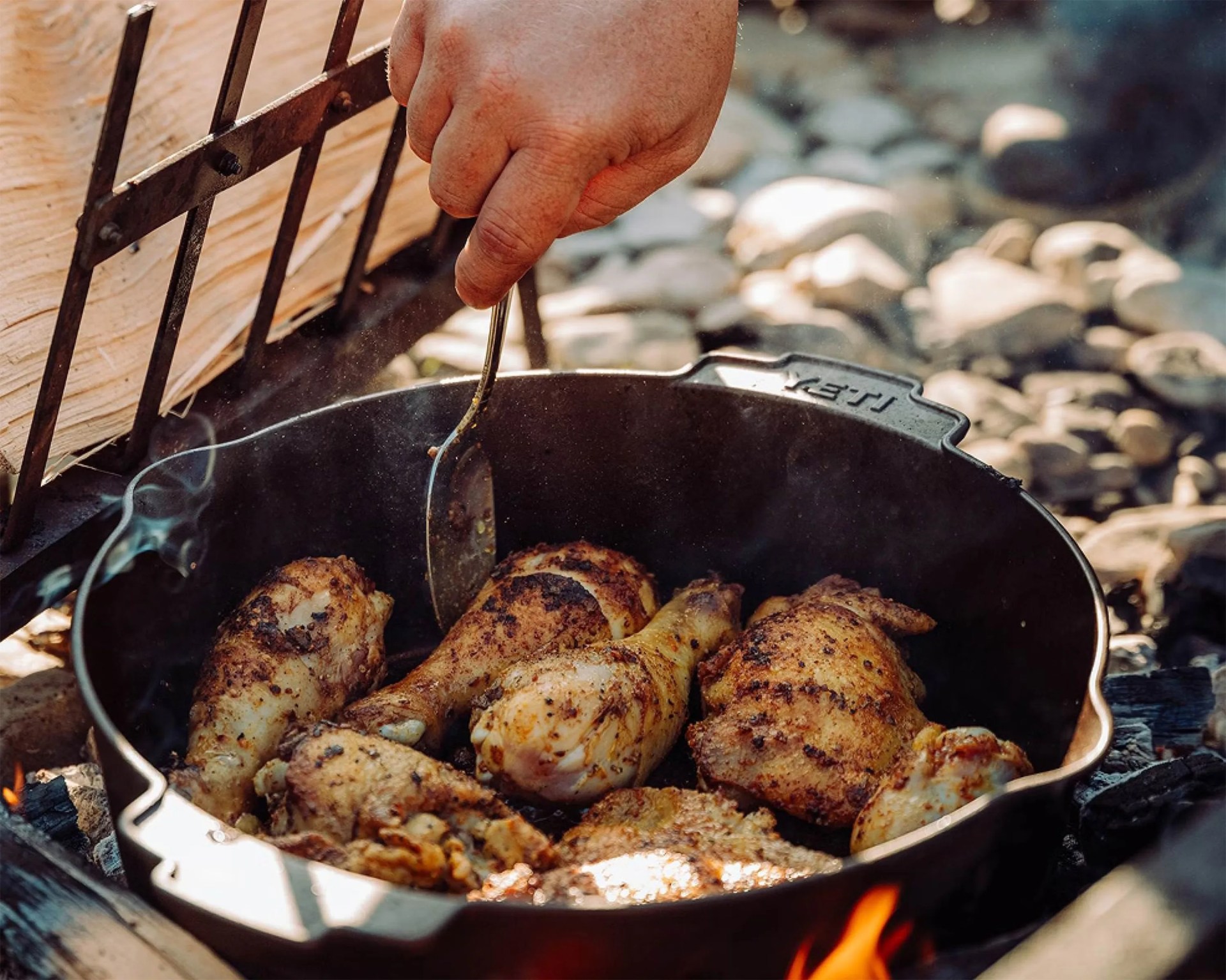 Cast iron skillet with seasoned chicken drumsticks and thighs cooking over an open fire, hand holding a spatula.