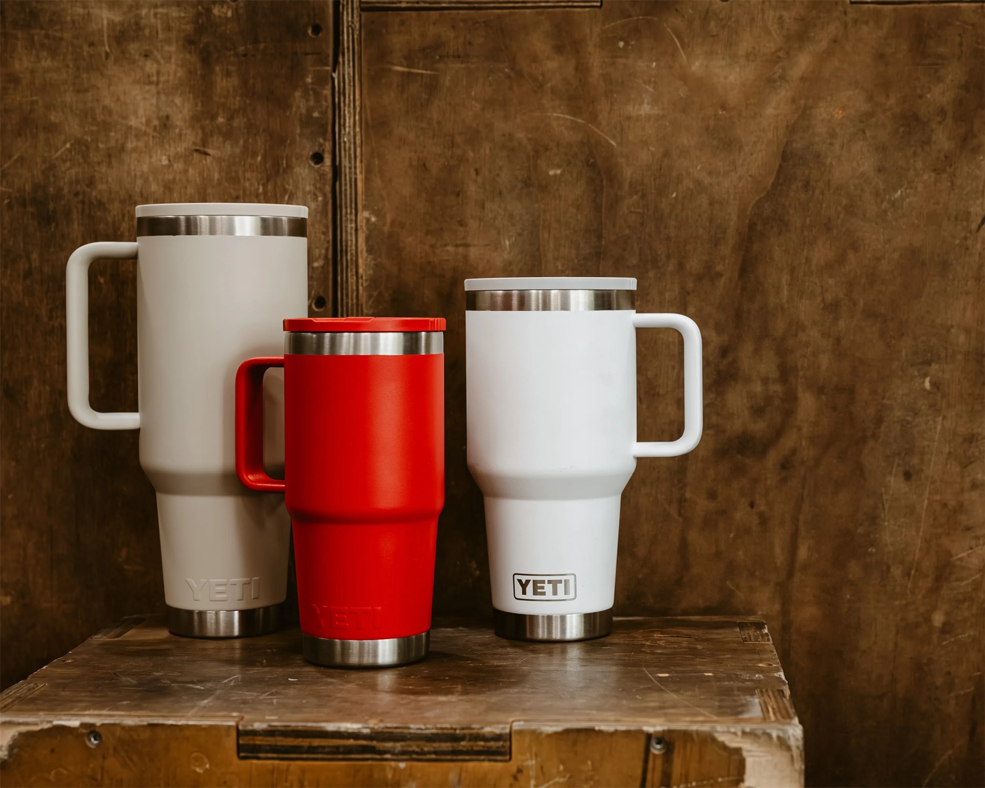 Three YETI insulated mugs in white, red, and light gray with handles, placed on a wooden surface against a rustic wooden background.