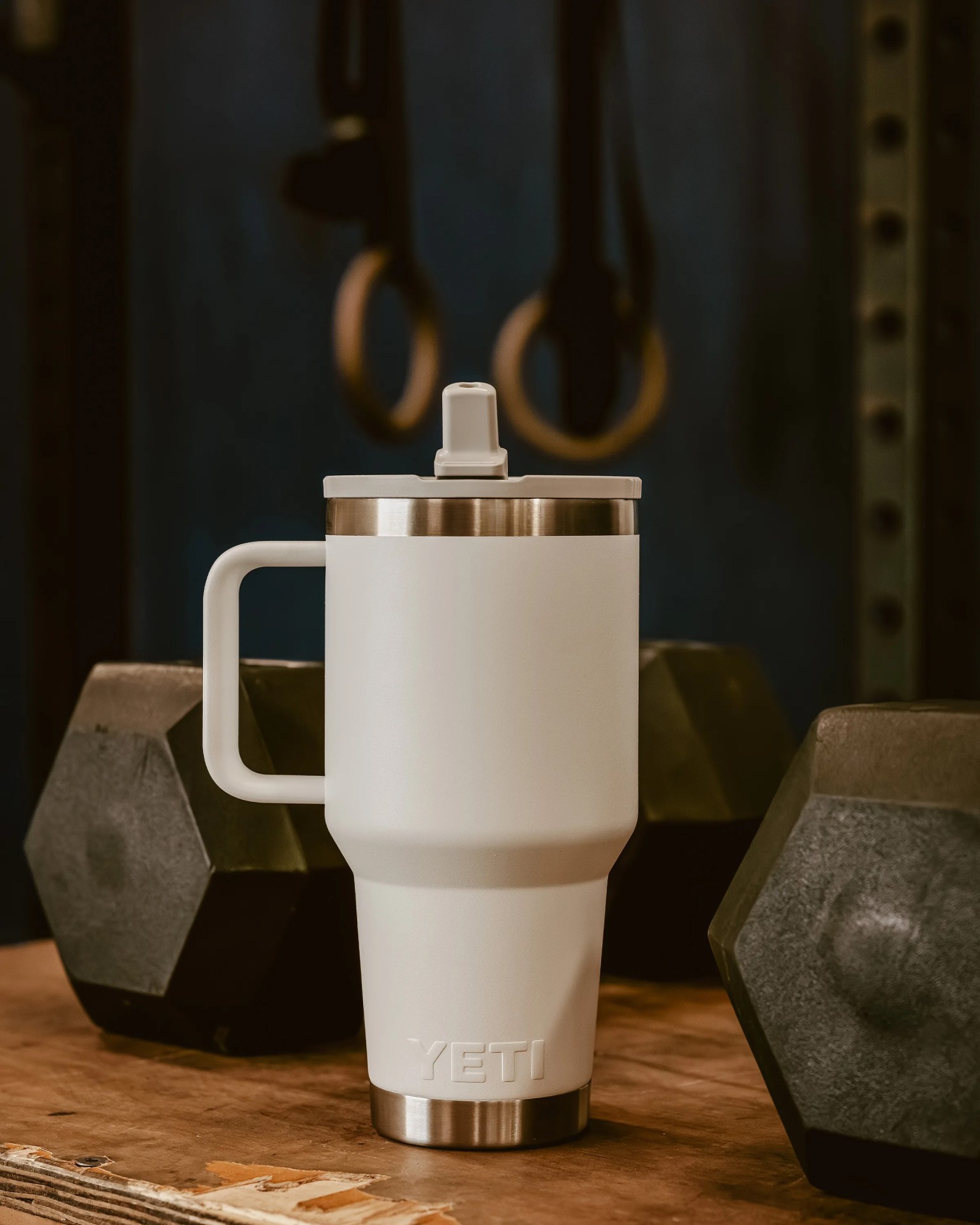 White YETI insulated tumbler with handle and lid on a wooden surface, surrounded by black hex dumbbells.