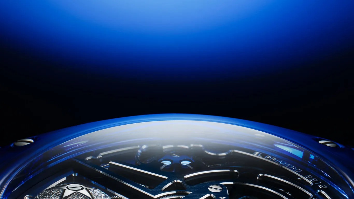 Close-up of a blue watch bezel with visible "EL PRIMERO 8612" engraving and metallic watch hands under a curved crystal.