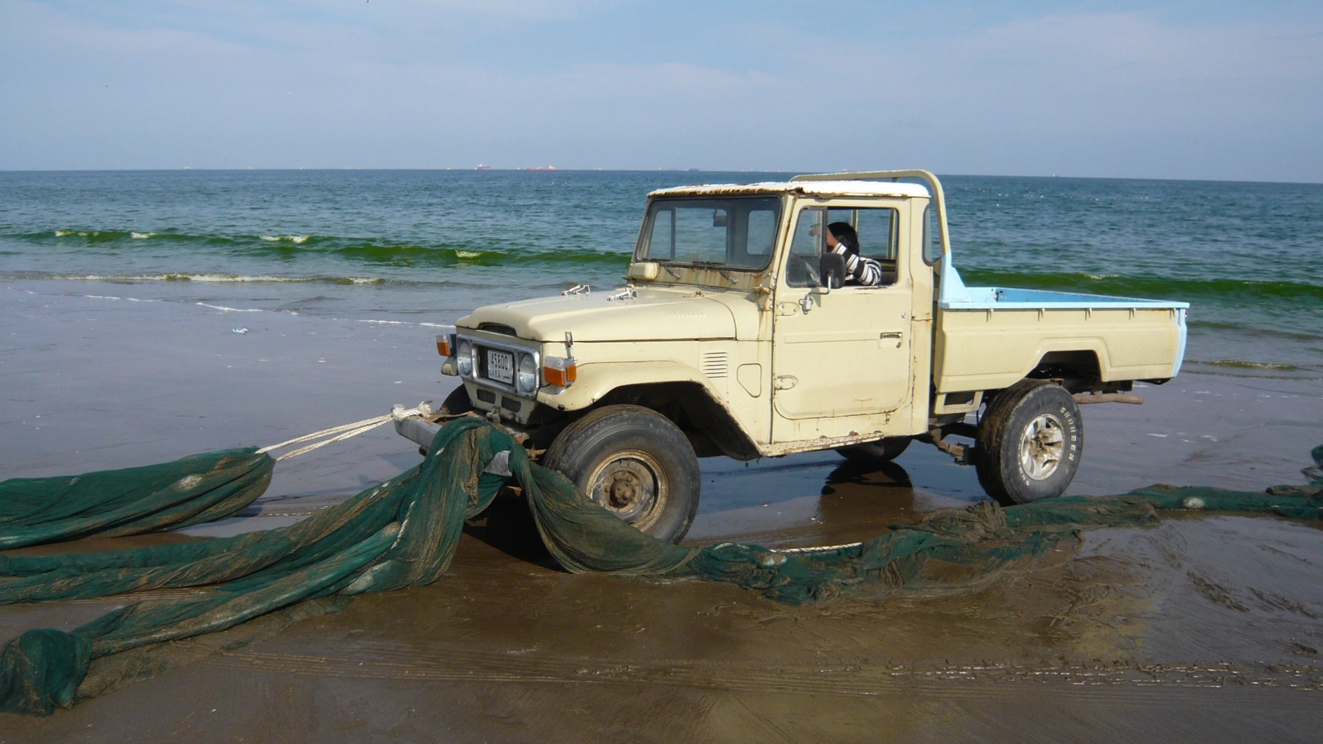 Beige vintage pickup truck on a wet sandy beach pulling green fishing nets with ocean waves in the background.
