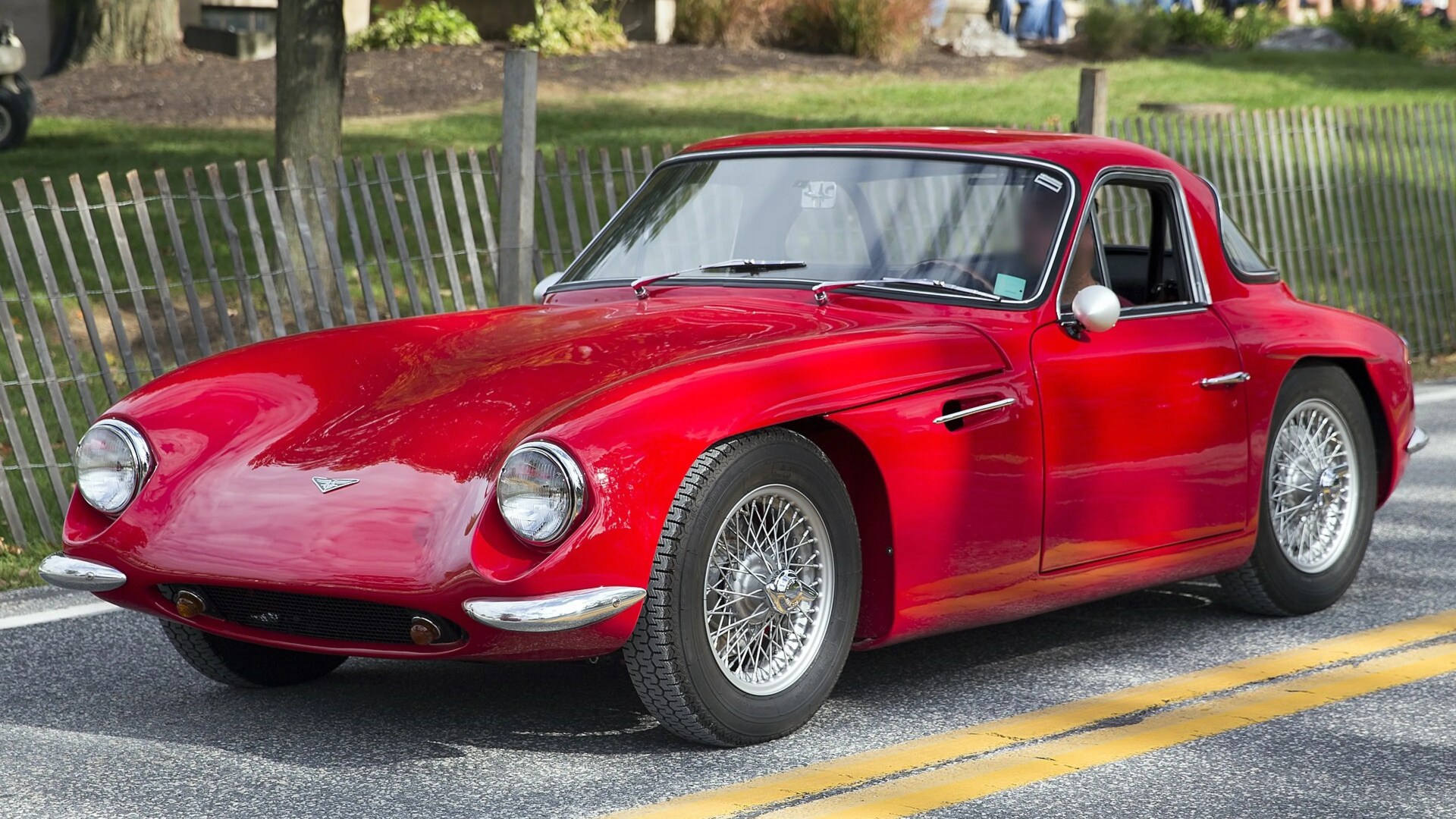Red vintage sports car with chrome wire-spoke wheels parked on a road with a wooden fence in the background.
