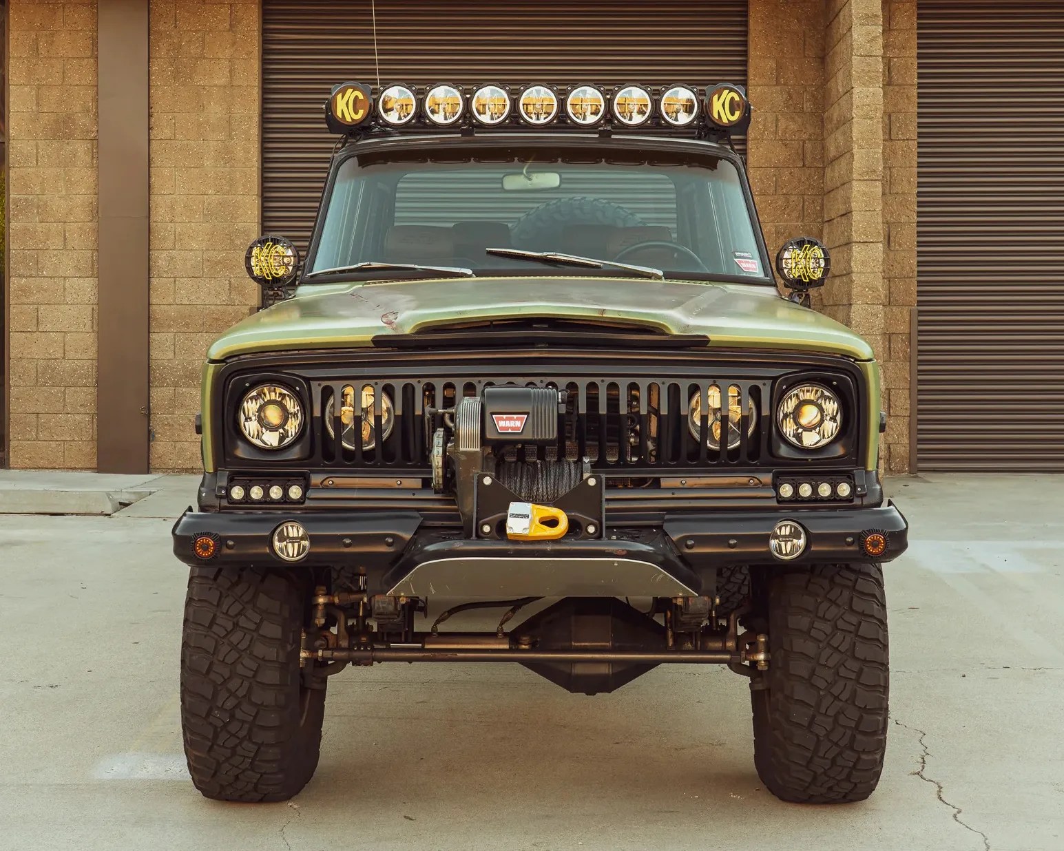 Front view of a green off-road vehicle with large tires, multiple KC lights on the roof and sides, and a WARN winch mounted on the bumper.