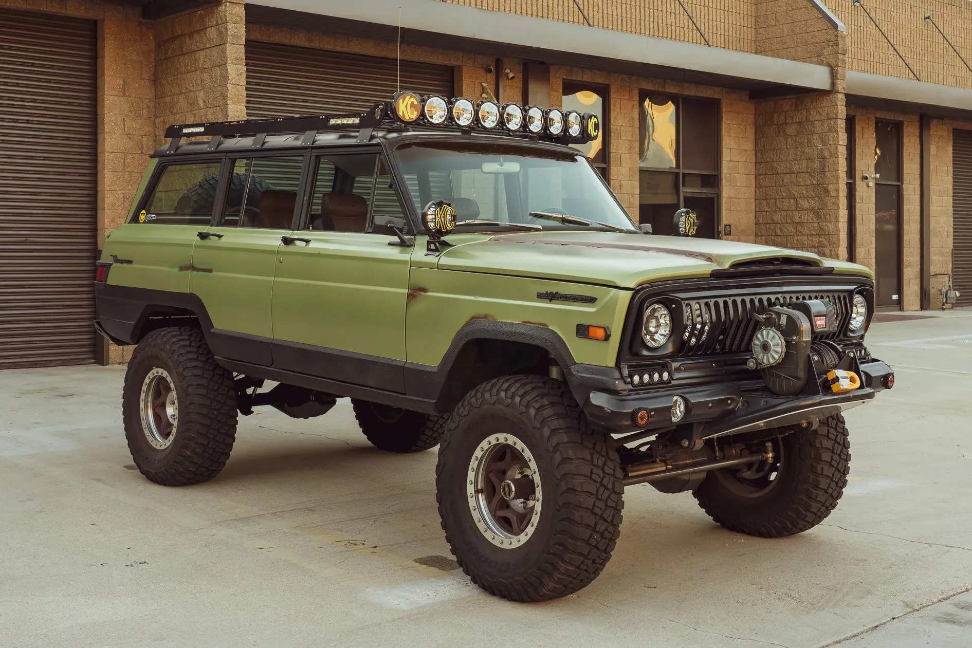 Green vintage off-road SUV with large tires, roof rack, multiple KC lights, and a front winch parked in front of a building.