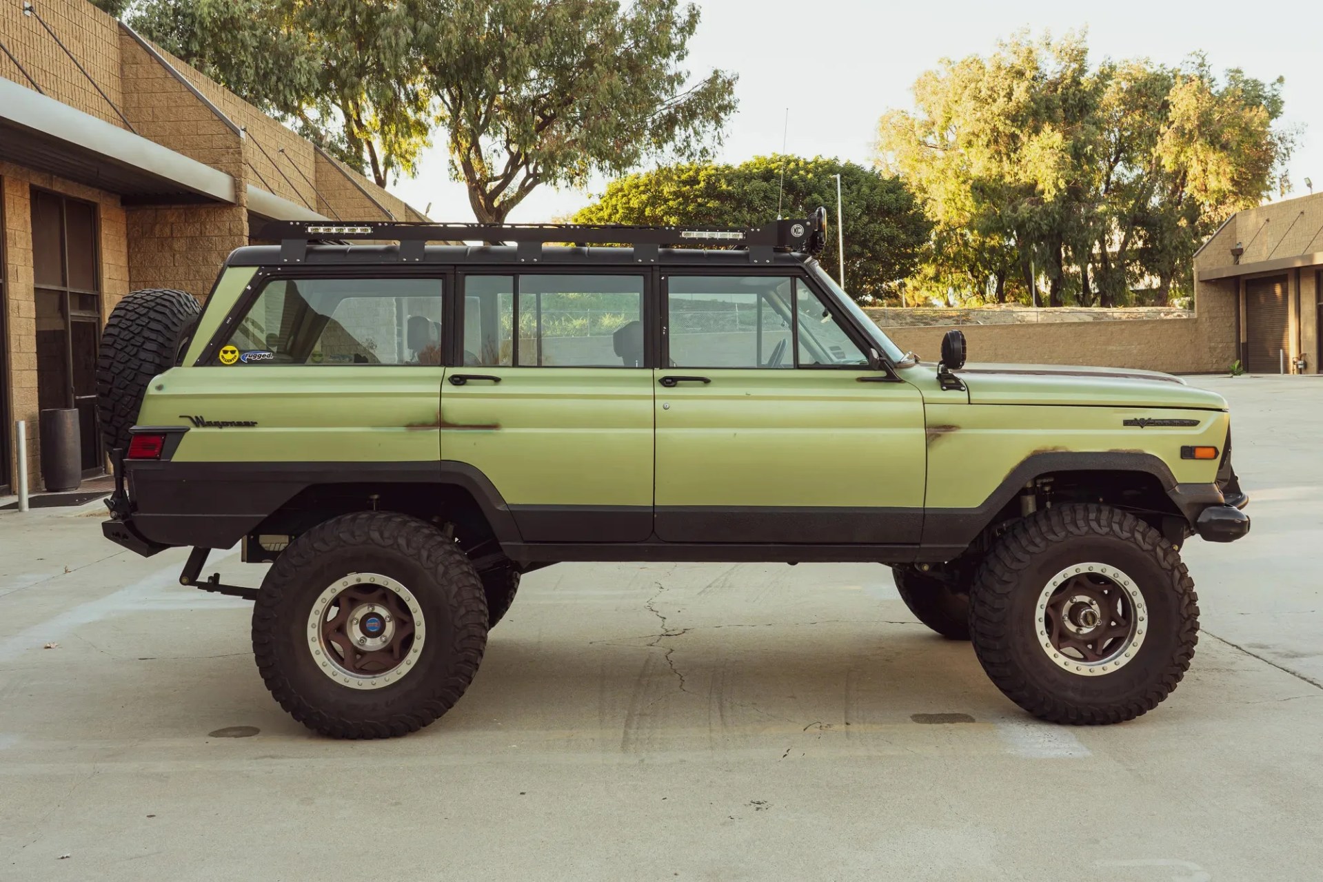 Light green Jeep Wagoneer with large off-road tires and black roof rack parked on concrete.