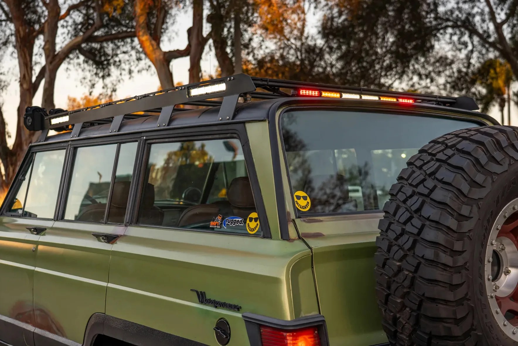 Green Jeep Wagoneer with roof rack and large off-road spare tire mounted on the rear.