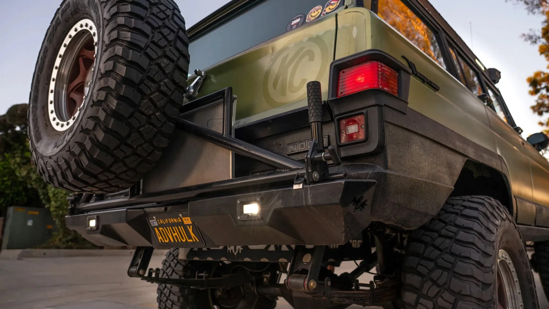 Rear view of an olive green off-road vehicle with large tires, a spare tire mounted on the back, and a California license plate reading "ADVHULK.