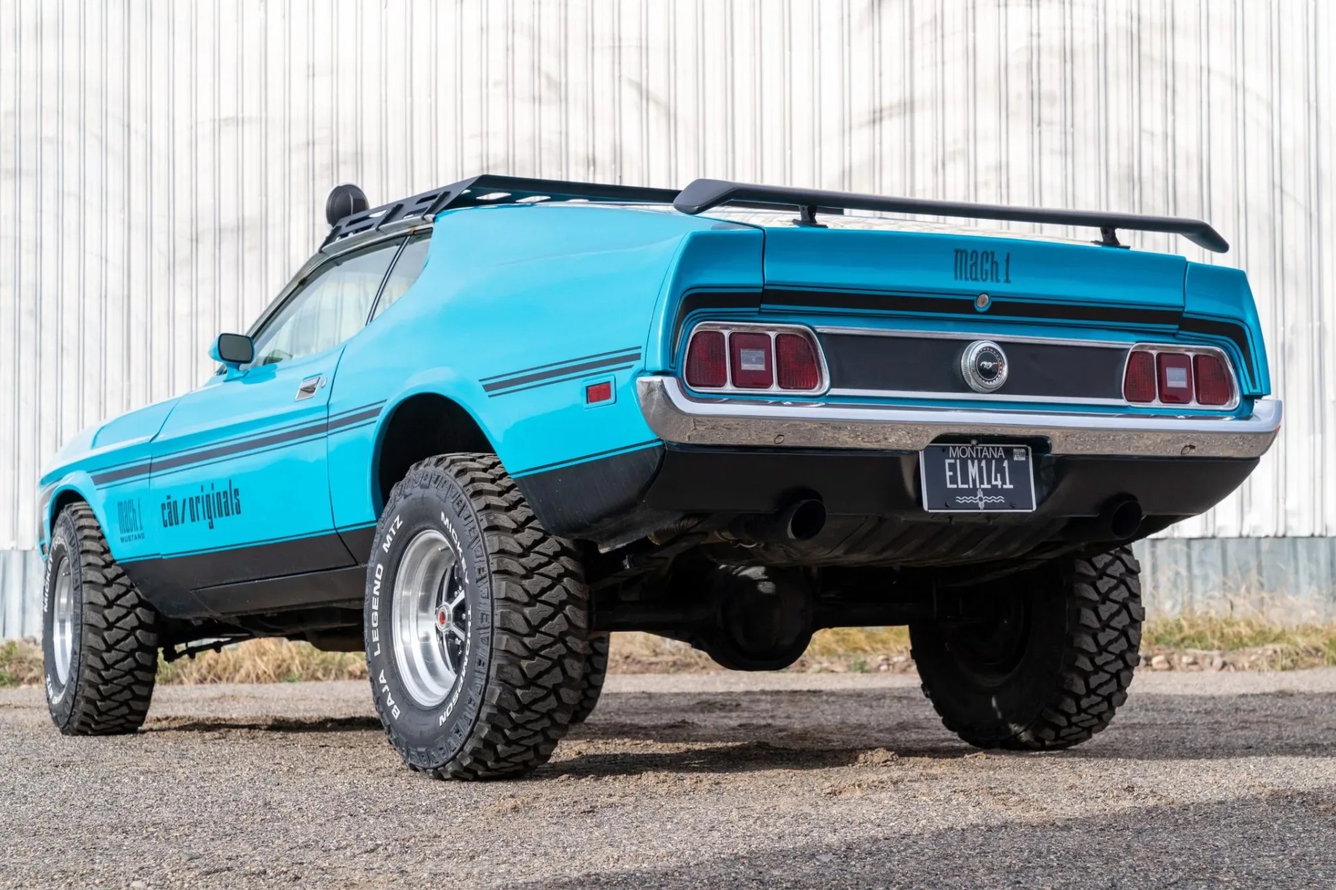 Bright blue Ford Mustang Mach 1 with off-road tires and roof rack parked on gravel in front of a corrugated metal wall.