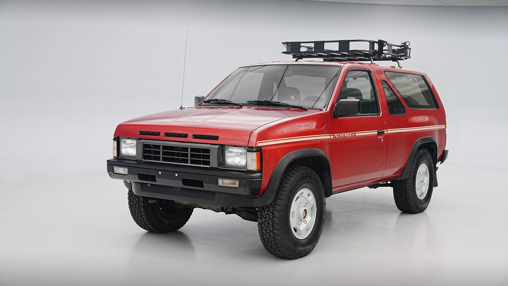 Red two-door Nissan Pathfinder SUV with roof rack and off-road tires in a white studio.