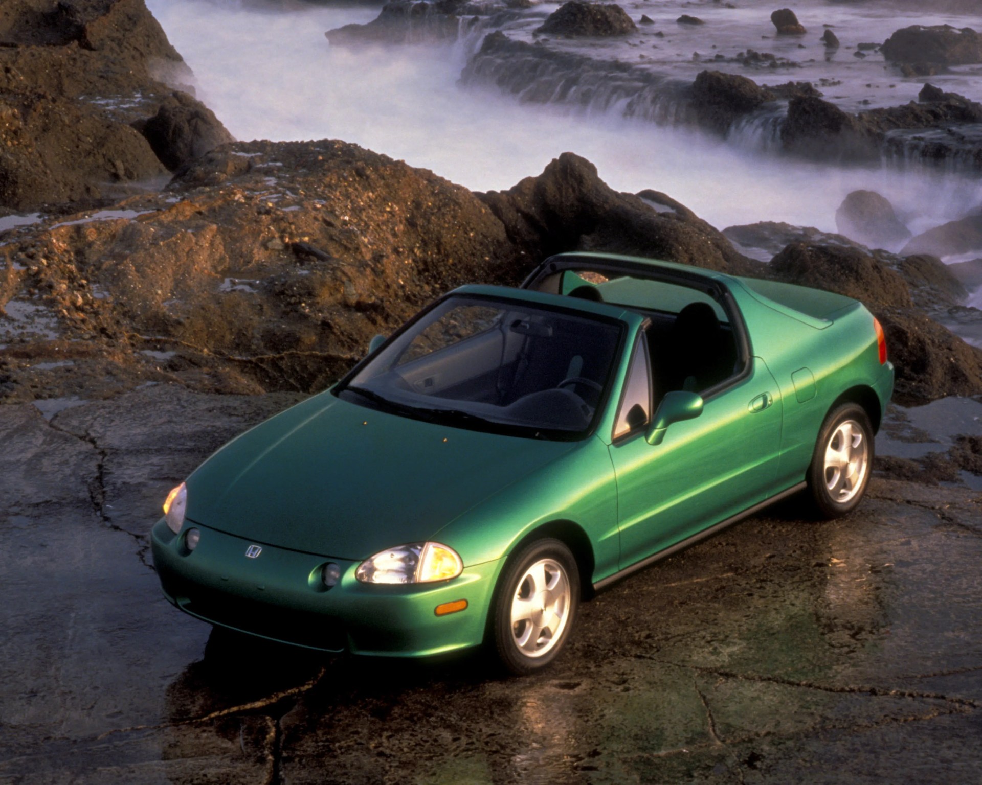 Green two-door Honda convertible parked on rocky shore with misty water in the background.