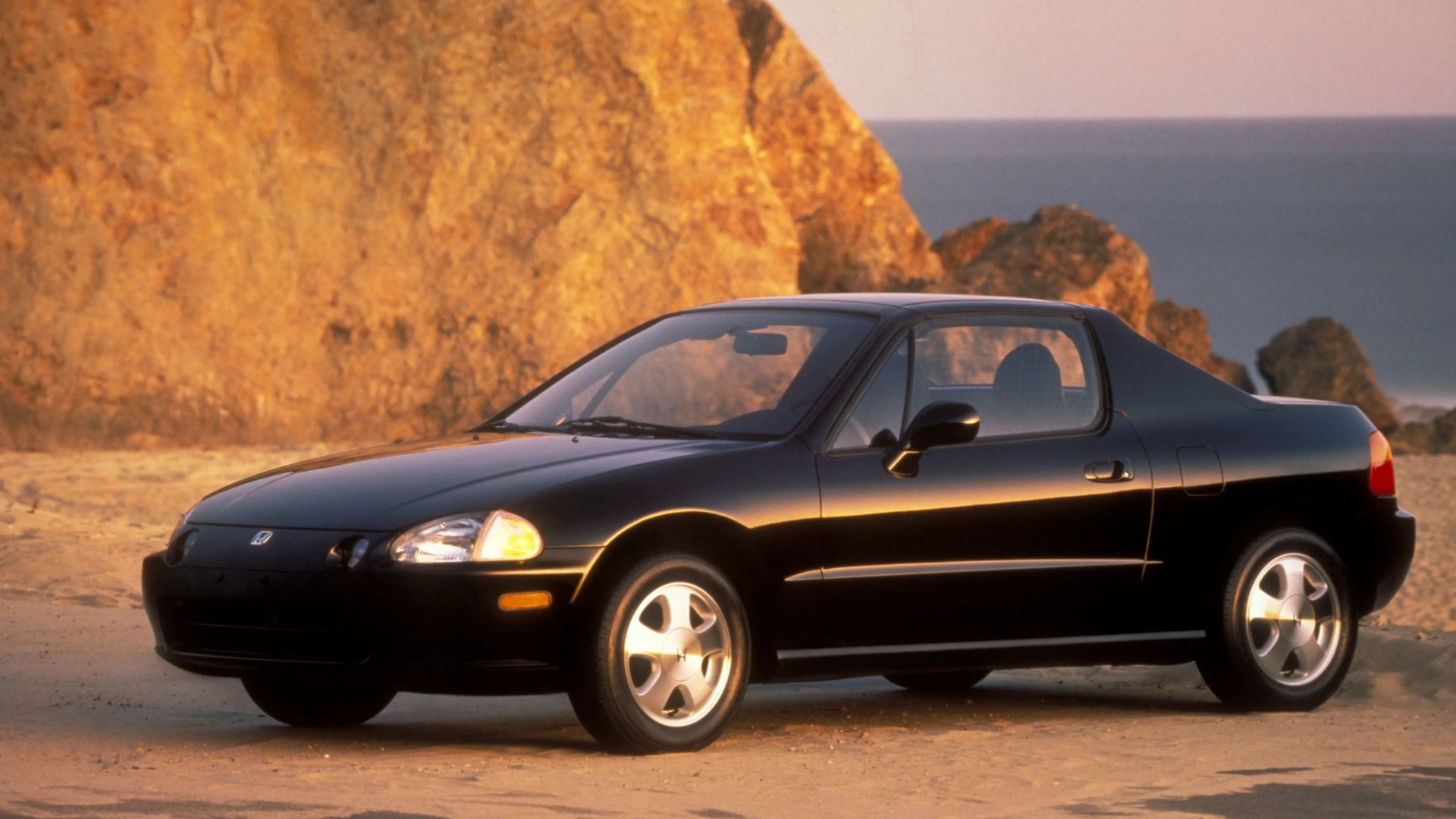Black two-door Honda CR-X coupe parked on sandy ground with rocky cliffs and ocean in background.