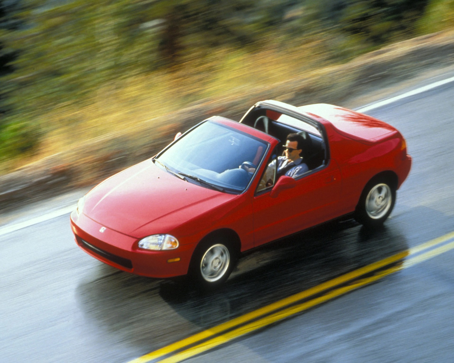 Red two-seater convertible sports car driving on a wet road with a person wearing sunglasses behind the wheel.