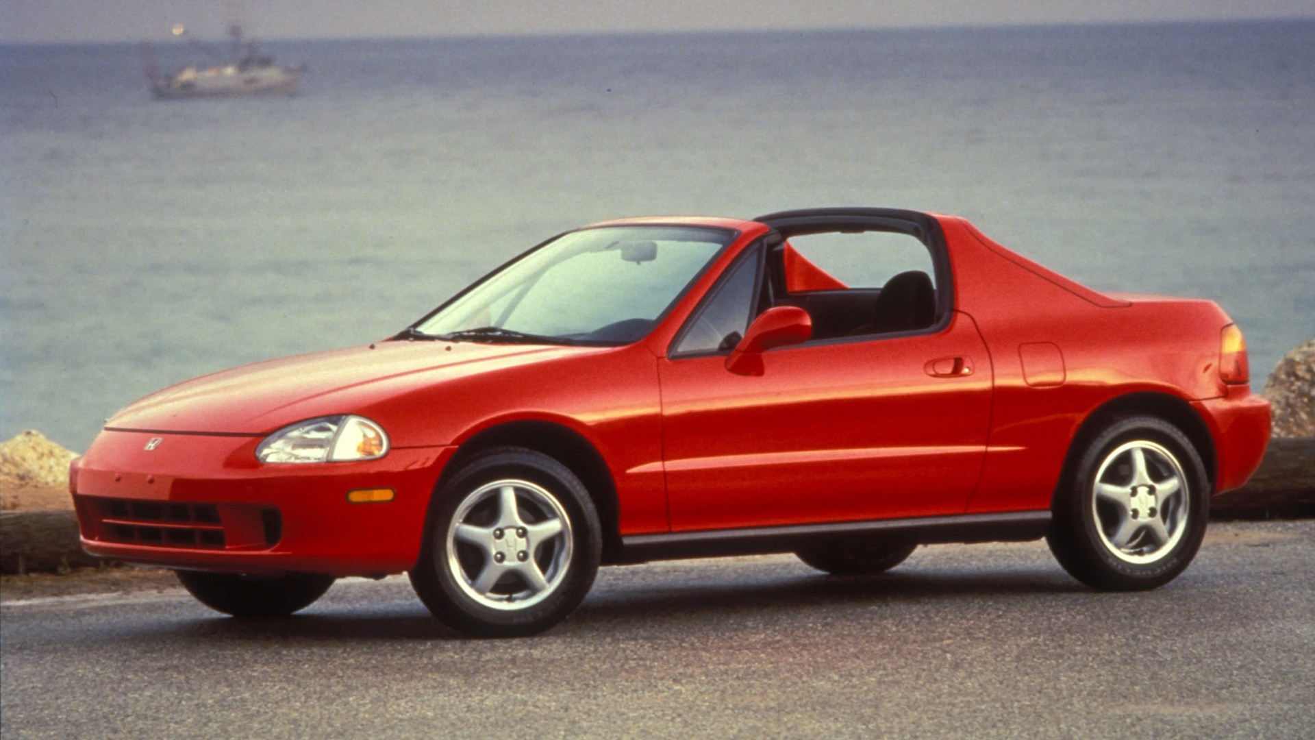 Red two-door Honda convertible parked on a road with ocean in the background.