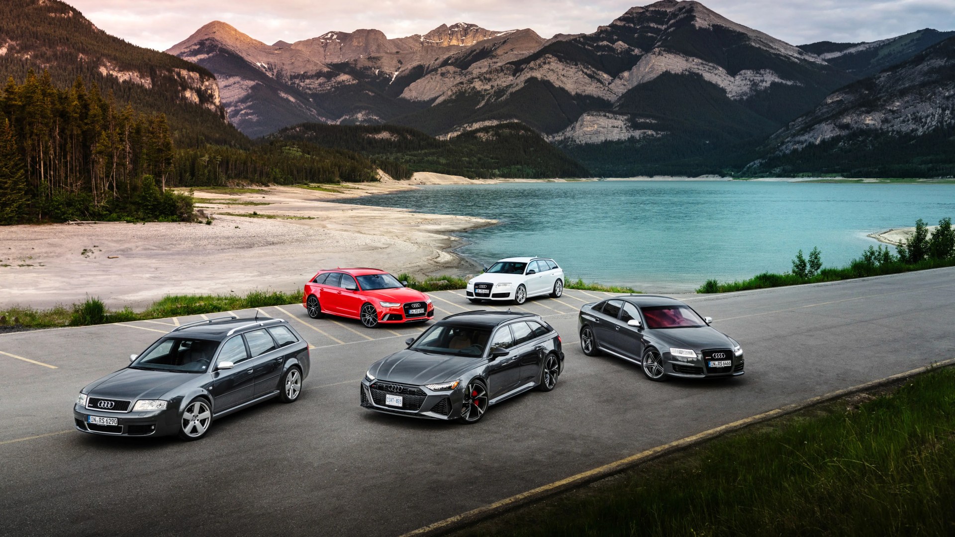 Five Audi RS models in various colors parked on a lakeside road with mountains in the background.