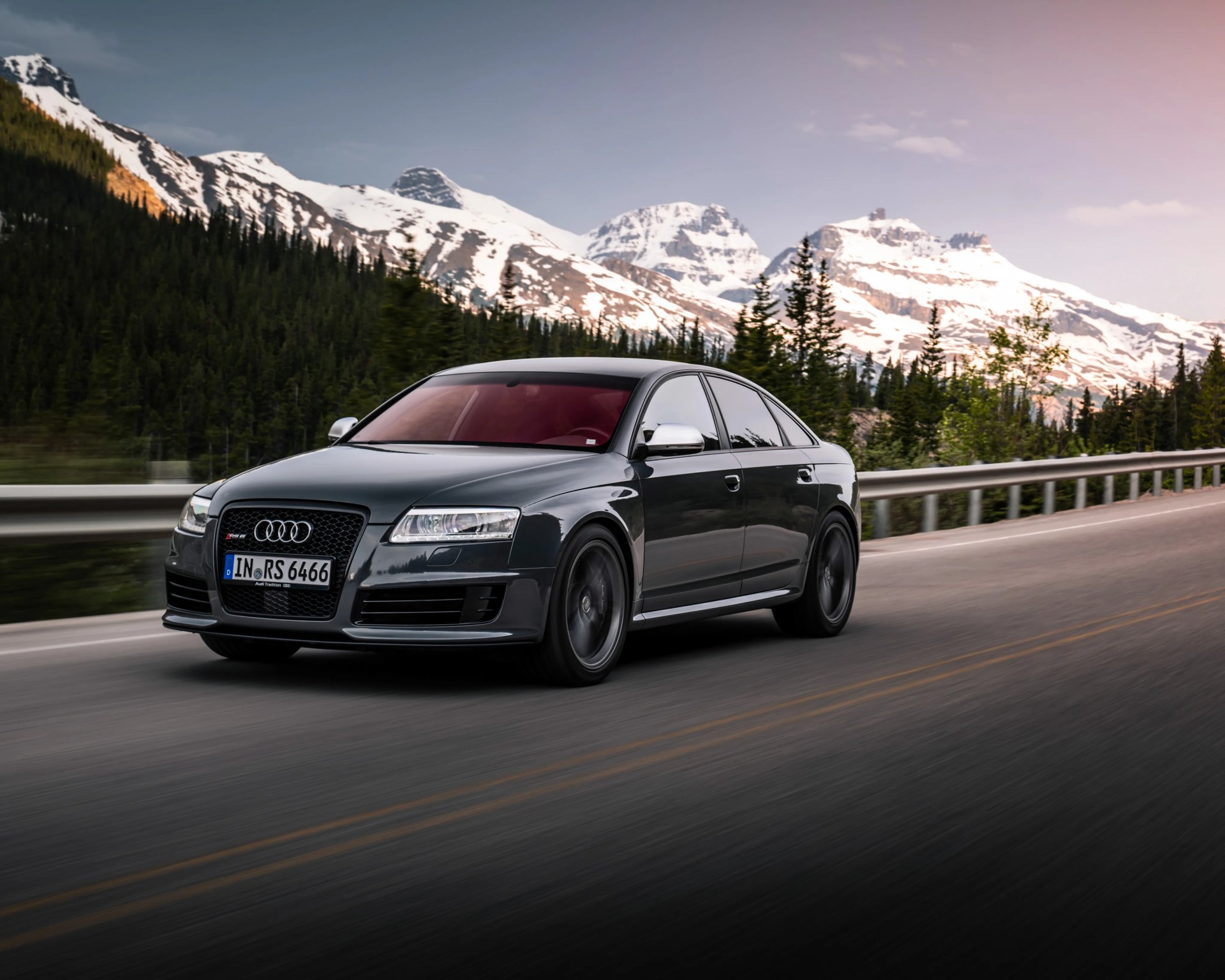 Dark gray Audi RS6 sedan driving on a mountain road with snow-capped peaks and pine trees in the background.